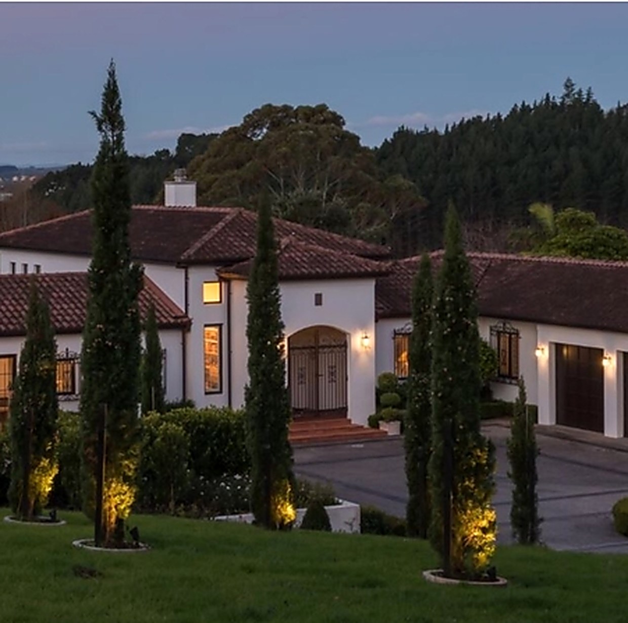 A Spanish-style Auckland home at dusk, with clipped hedging, uplit cypresses and lawn running down to the drive